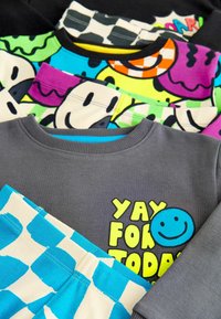Grå sweatshirts med teksten "YAY FOR TODAY" og et smiley-ansigt, lagd med farverige mønstrede toppe og blå polkaprikkede shorts. Blødt bomuldsstof.