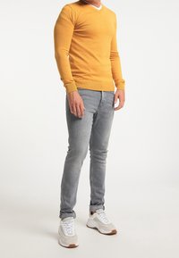 Gul V-ringad tröja med långa ärmar, kombinerad med ljusgrå jeans och vita sneakers med bruna detaljer. Slät textur, åtsittande design.