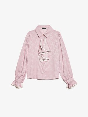 Blouse rose clair avec un devant volanté, tissu à motifs de nœuds blancs, et manches longues avec des poignets à volants. Fermeture à boutons sur le devant.