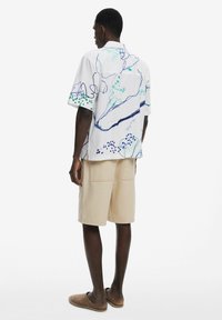Camicia a maniche corte con base bianca, caratterizzata da motivi astratti blu e verdi, abbinata a pantaloni corti beige e scarpe slip-on marroni.