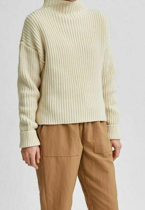 Personne portant un pull en tricot côtelé couleur crème avec des manches longues et un col montant, associé à un pantalon ample marron clair avec de grandes poches avant.