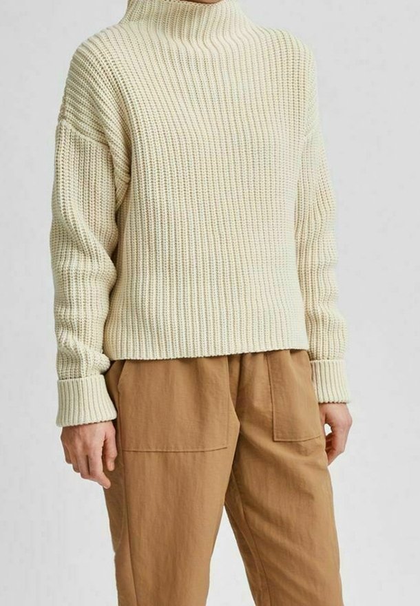 Personne portant un pull en tricot côtelé couleur crème avec des manches longues et un col montant, associé à un pantalon ample marron clair avec de grandes poches avant.