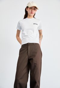 T-shirt branco justo com o logótipo "DICKIES" no peito, combinado com calças castanhas largas. O modelo usa um boné bege claro.