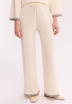 Kremowe spodnie o szerokich nogawkach ze ściągaczem w talii, noszone z dopasowanym topem i beżowymi butami z kwadratowym noskiem na jednolitym tle.