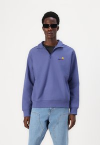 Purple zip-up sweatshirt with a high collar, featuring a small embroidered logo. Worn over a black textured shirt with blue denim jeans.