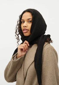 Sort uldscarf draperet over hovedet, med en blød struktur. Modellen har en lys brun frakke på, med krøllet hår der rammer ansigtet.