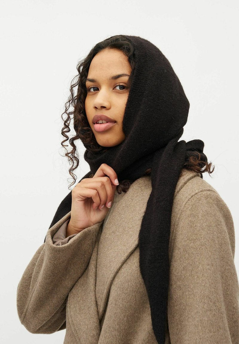 Sort uldscarf draperet over hovedet, med en blød struktur. Modellen har en lys brun frakke på, med krøllet hår der rammer ansigtet.