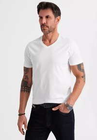 T-shirt bianco a maniche corte con scollo a V, realizzato in cotone, indossato con jeans scuri. Presenta tatuaggi visibili sulle braccia e un orologio d'argento al polso.
