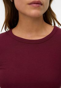 Femme portant un haut bordeaux côtelé avec un col rond et des boucles d'oreilles en or, montrant le bas du visage et le cou.