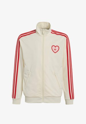 Kermanvärinen vetoketjullinen urheilutakki, jossa punaiset kolmet raidat hihoissa ja punainen sydämenmuotoinen logo vasemmassa rinnassa.