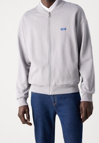 Mand iført en lys grå zip-up jakke med et blåt "Hugo" logo på brystet og blå jeans, der står foran en ensfarvet baggrund.