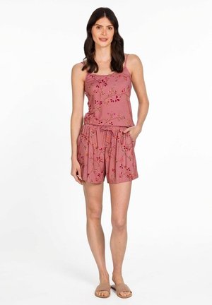 Vrouw draagt een roze, mouwloze top met bloemenmotief en bijpassende korte broek, met haar hand in de zak, staand tegen een witte achtergrond.