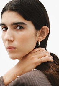 Pendientes de aro dorados con un diseño puntiagudo y un anillo minimalista a juego. La modelo tiene el cabello lacio y oscuro, y una expresión neutral.
