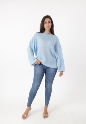 Maglione azzurro chiaro a maglia larga con maniche ampie, abbinato a jeans skinny blu scuro e sandali con tacco trasparenti, su uno sfondo semplice.