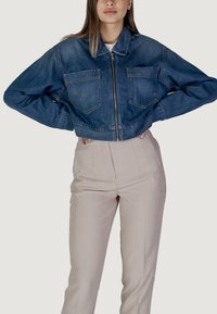 Giacca in denim cropped con zip frontale, due tasche sul petto e un lavaggio blu scuro. Abbinata a pantaloni su misura di colore chiaro.