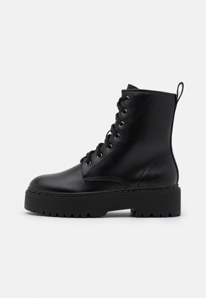 Schwarzer Leder-Schnürstiefel mit dicker Profilsohle und Zuglasche hinten, von der Außenseite auf weißem Hintergrund gezeigt.