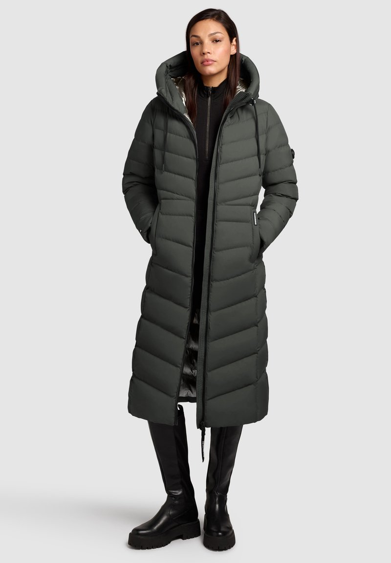Long manteau en duvet vert foncé, doté d'une capuche, d'un design matelassé en chevrons, d'une fermeture éclair avant et de poches latérales. Associé à des bottes noires jusqu'aux genoux.