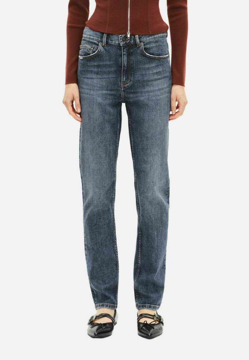 Jeans de mezclilla azul con un corte recto, cinco bolsillos y detalles de costura sutiles. Combinados con bailarinas negras sobre un fondo simple.