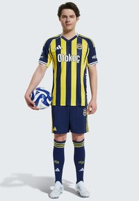 Futbalový dres Fenerbahçe v žltých a námorníckych pruhoch, s bielymi akcentmi, s hráčom držiacim modro-biely loptu a oblečeným v bielych kopačkách.