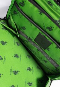 Vihreä sisustusmateriaali laukussa, jossa on toistuvia mustia logopainatuksia. Mukana nahkalamina ja useita lokeroita järjestämiseksi.