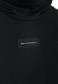 Top negro de cuello alto hecho de una tela suave y elástica, que presenta una etiqueta negra rectangular con texto blanco cerca del escote.