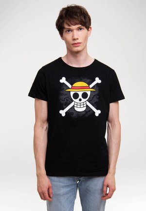Young man wearing a black t-shirt with a white skull and crossbones design featuring a yellow and red straw hat, paired with light blue jeans.