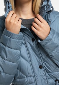 Pikowana kurtka w jasnoniebieskim materiale, z kapturem i zapięciem na guziki. Model trzyma kołnierz zamknięty.