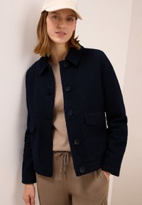 Kvinna som bär en beige keps, marinblå texturerad jacka med knappar, beige topp och ljusbruna byxor med dragsko, stående mot en ljus vägg.