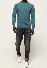 Felpa teal con colletto rotondo e tessuto texturizzato, abbinata a jeans scuri e sneakers bianche. Dettaglio sulle spalle e logo distintivo.