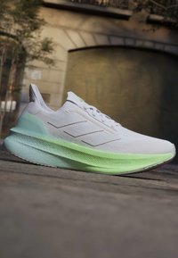 Chaussure de course blanche et vert clair avec trois bandes latérales, semelle épaisse et rembourrée, et fermeture à lacets sur une surface extérieure pavée.