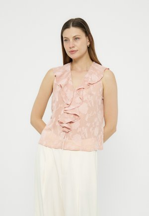 Kobieta z długimi brązowymi włosami, ubrana w bezrękawnikową bluzkę w bladym różowym kolorze z falbanami oraz białe, szerokie spodnie na jednolitym tle.