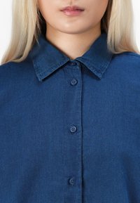 Blondýna v tmavě modré košili s límečkem, zapnuté až ke krku, zobrazená od brady po hruď na bílém pozadí.