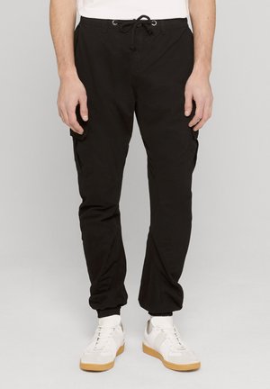 Pantaloni cargo jogger neri con vita regolabile e polsini elasticizzati, indossati con sneakers bianche e calzini su una persona in piedi.