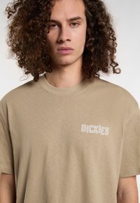 Tan short-sleeve t-shirt made of soft cotton. Features a textured white "DICKIES" logo on the left chest area. Classic crew neck design.