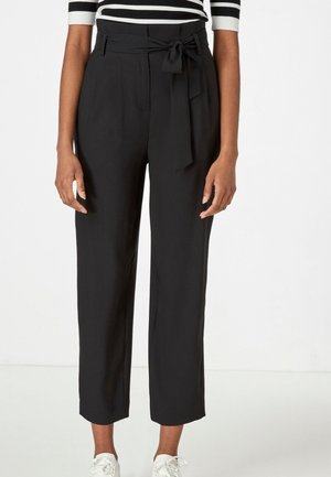 Pantalon noir taille haute avec une ceinture à nouer, présentant une coupe large et une texture lisse, associé à des baskets blanches.
