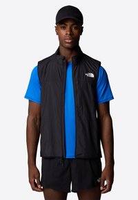 Black sleeveless vest with a high collar, lightweight material, paired with a blue short-sleeve shirt, complemented by a black cap.