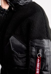 Schwarze Fleecejacke mit Lederakzenten, Hohem Kragen, Rippbündchen und mehreren Taschen. Verfügt über einen roten Reißverschluss-Anhänger mit der Aufschrift "REMOVE BEFORE."