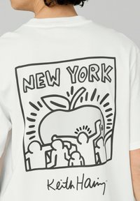 T-shirt blanc présentant des dessins en ligne noire de personnes et d'une pomme, avec le texte "NEW YORK" au-dessus et la signature "Keith Haring" en dessous. Tissu en coton confortable.