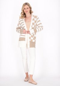 Cardigan checkered beige e bianco con maniche lunghe, apertura frontale e due tasche frontali, abbinato a pantaloni bianchi e scarpe crema.