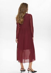 Rochie lungă bordeaux cu mâneci lungi transparente, buline texturate și o fustă fluidă. Asortată cu pantofi cu toc înalt negri.