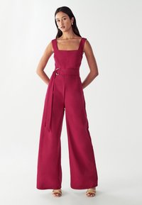 Bordeaux jumpsuit met wijde pijpen en een vierkante halslijn. Aansluitende taille met een bijpassende riem met een zilveren gesp. Zachte, lichte stof.