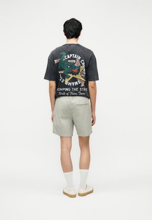 Osoba ubrana w czarny t-shirt z zielonym smokiem i białym napisem, szare krótkie spodenki, białe skarpetki i białe trampki, stojąca tyłem.