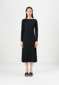 Robe noire à manches longues avec un col bateau, un buste ajusté et une jupe fluide. Associée avec des sandales noires à enfiler. Texture du tissu lisse.
