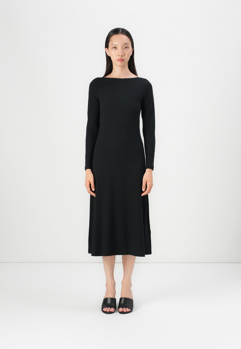 Robe noire à manches longues avec un col bateau, un buste ajusté et une jupe fluide. Associée avec des sandales noires à enfiler. Texture du tissu lisse.
