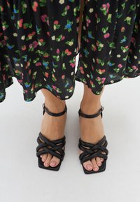 Des sandales noires à lanières avec un bout carré, arborant des brides en cuir croisées et une boucle à la cheville. Portées avec une jupe à motifs floraux.