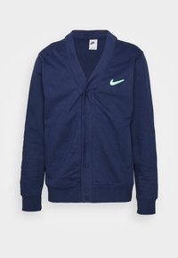 Cardigan Nike bleu marine avec boutons-pression, col en V, manches longues, poignets côtelés et logo Swoosh vert de Nike sur la poitrine gauche.