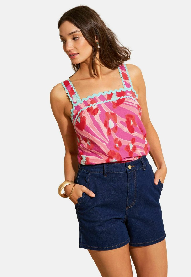 Vrouw draagt een roze bloemenmouwloze top met blauwe gegolfde rand en hooggetailleerde donkere denimshorts, handen in de zakken.