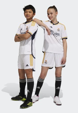 Camisetas de fútbol blancas con acentos en azul marino y dorado, combinadas con pantalones cortos y medias negras a rayas. Los jugadores llevan zapatos de fútbol en blanco y negro.
