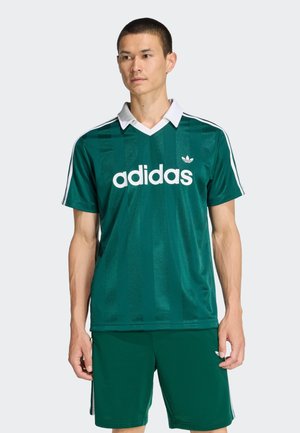 Maglia sportiva adidas verde con colletto bianco, caratterizzata da strisce verticali e grande logo bianco sul davanti, indossata con pantaloncini abbinati.