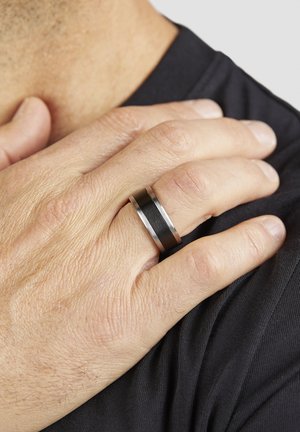 Nahaufnahme einer Hand, die auf einer Schulter ruht, mit einem breiten schwarz-silbernen Ring am Ringfinger vor einem schwarzen Hemdhintergrund.